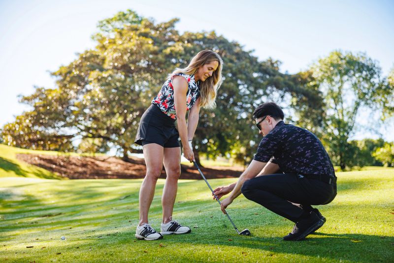Young Caucasian female golfer receiving guidance from male instructor on golf course with lush greenery and bright sunlight, both concentrating on improving swing technique.
