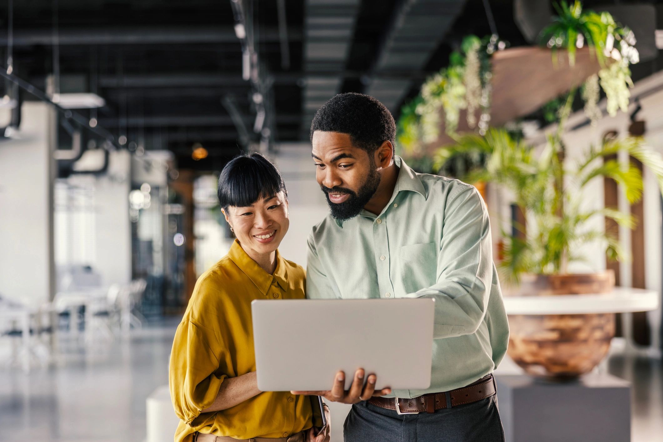 Two coworkers smiling while looking at a laptop in a modern office.