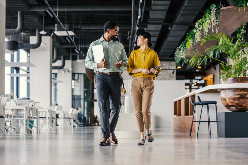 Two diverse business people walking through a contemporary office space, holding a laptop and tablet, engaging in a focused discussion about their work and collaboration