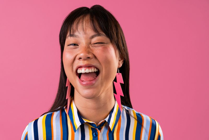 Beautiful chinese woman portrait on colorful background. Cheerful young asian woman winking and laughing with mouth open, wearing colorful striped shirt and pink lightning bolt earrings