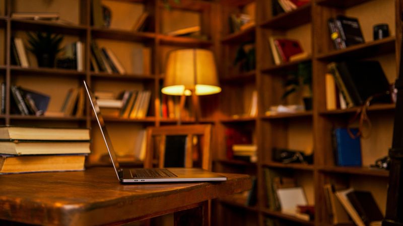 A warm, inviting study space featuring floor-to-ceiling bookshelves filled with various books, a vintage lamp, and a modern laptop open on a rustic wooden table