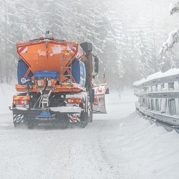 Snowplow truck clearing and salting a snowy road in a forest.
