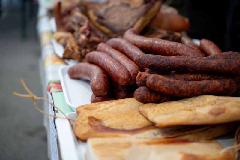 A selection of smoked sausages and slabs of cured pork are displayed on a table at an outdoor market. The rich textures and deep, reddish hues highlight traditional meat preparation methods. The arrangement evokes a rustic, artisanal food culture with a focus on quality and flavor.