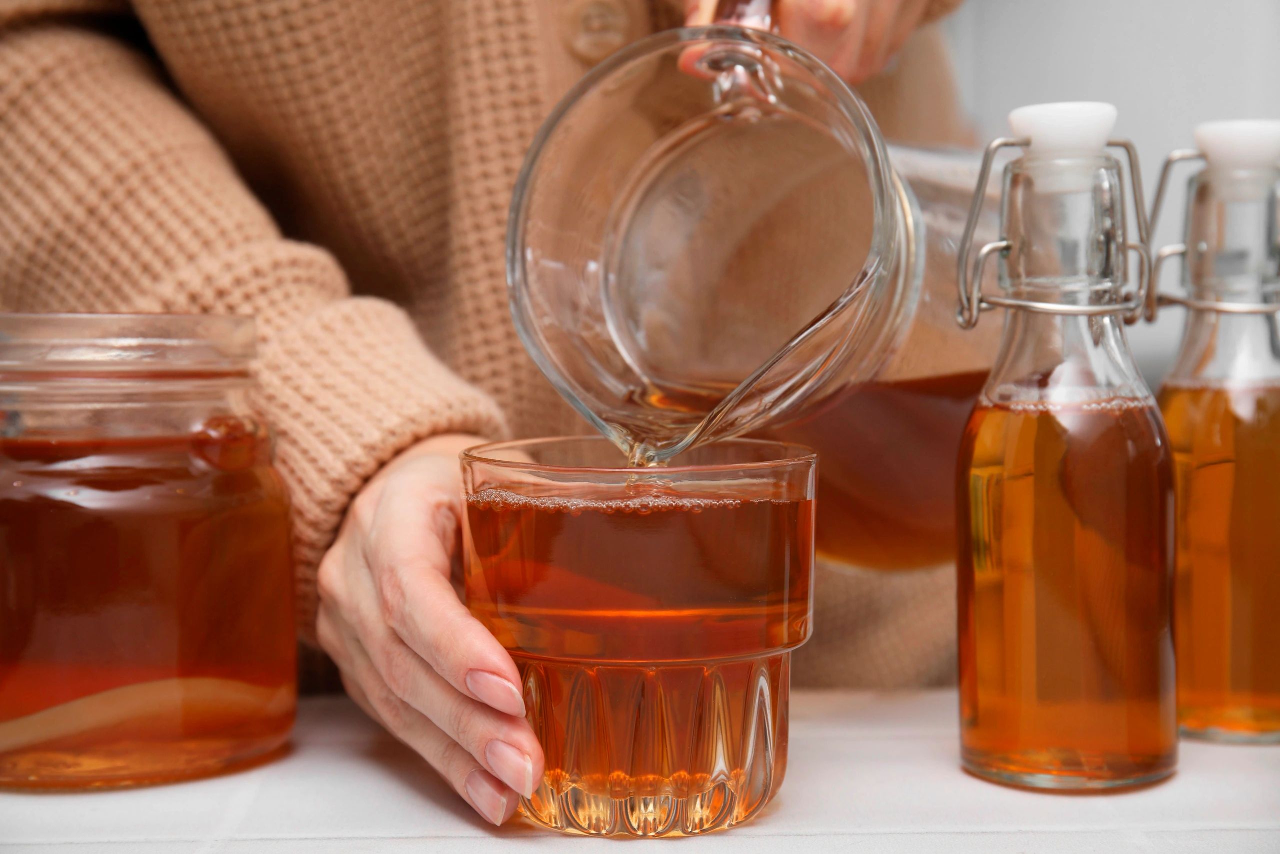 Woman pouring fermented kombucha from jug into glass at white table, closeup
