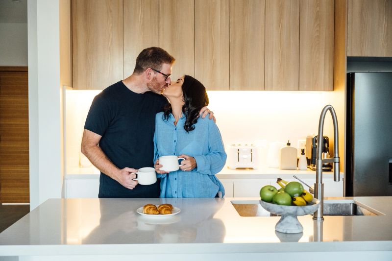 Mid adult Australian couple standing in the modern kitchen behind the counter and drinking morning coffee or tea, expressing positivity and hugging