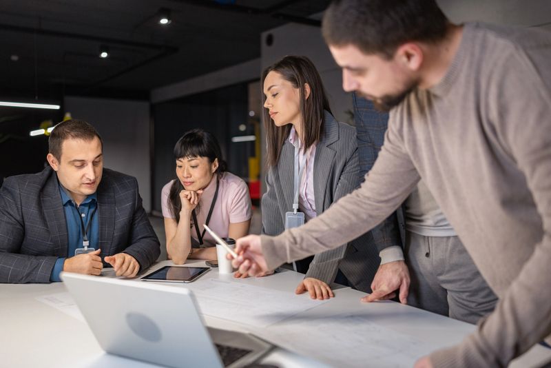 A group of multi-ethic colleagues gather around a table in a modern office to review blueprints and digital data, illuminated by focused overhead lighting.