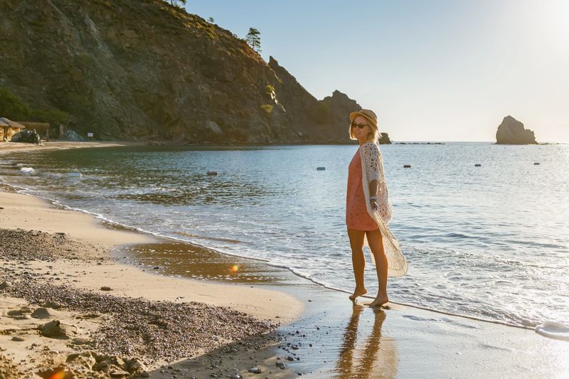 A medium shot of a stylish woman wearing a pink dress, a bohemian white lace cover-up, and a straw hat. She is standing barefoot on the sandy shoreline