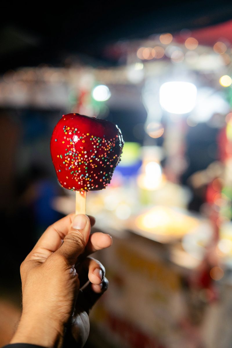 Hand holding a vibrant red candy apple covered in sprinkles at a festive night market