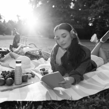 Young woman listening to music and reading outdoors on a picnic blanket.