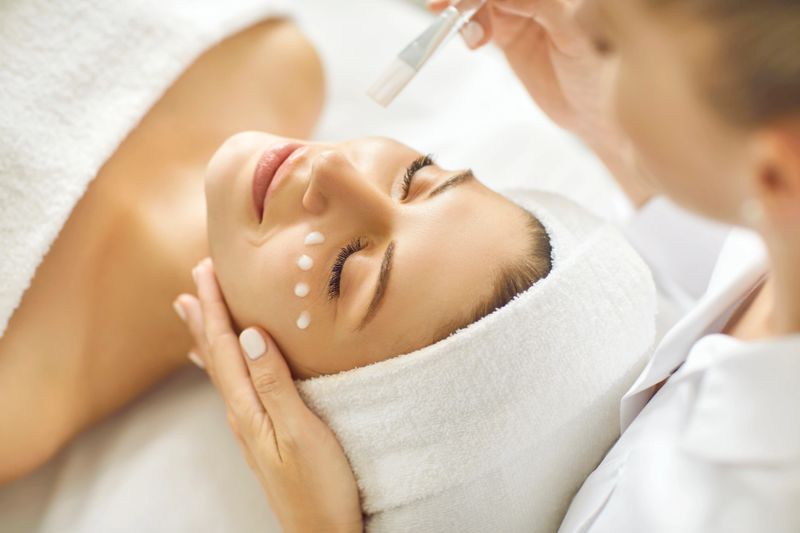Professional beautician doing facial treatment, applying cleansing, moisturizing, nourishing and rejuvenating mask on face of happy beautiful young woman at beauty parlor, spa salon or wellness center