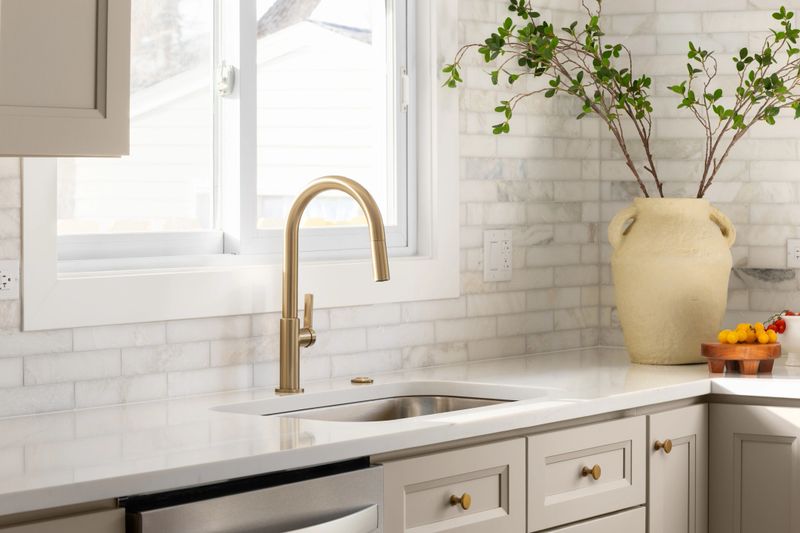 A gold kitchen faucet detail in a luxury kitchen with brown cabinets, marble subway tile backsplash, and decorations on the marble countertop.