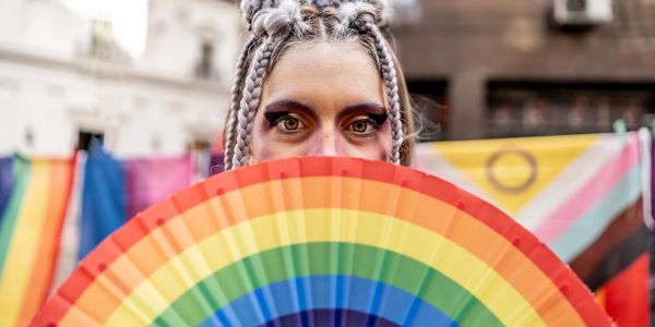 Person with braided hair holds a rainbow fan, colorful flags in background.