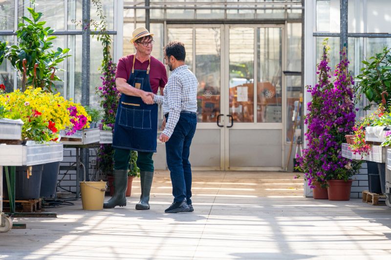 Mature Caucasian male gardener in greenhouse warmly engaging with Middle Eastern customer surrounded by vibrant plants and flowers, sharing plant care advice and gardening tips in a sunny greenhouse environment.