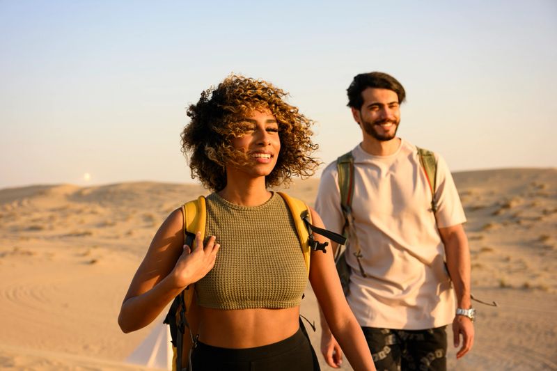 Young couple smiling as they hike across sunlit sand dunes with backpacks, enjoying a warm desert sunset and a carefree outdoor adventure together