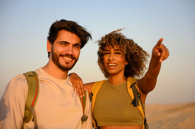 Young couple backpacking across sunlit desert dunes, smiling and pointing toward the horizon while enjoying an adventurous outdoor vacation together