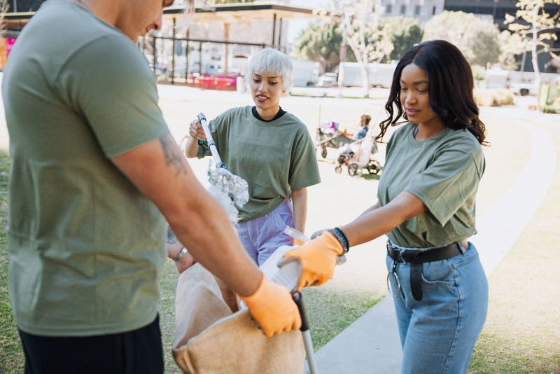 A small group of volunteers collaborates during a daytime cleanup event in a public park, removing trash and contributing to environmental preservation. The natural interactions, casual clothing, and urban background emphasize authenticity and community spirit. This visual story represents teamwork, environmental stewardship, and collective responsibility, making it suitable for editorial and commercial use related to sustainability, climate awareness, volunteerism, and community-driven change.