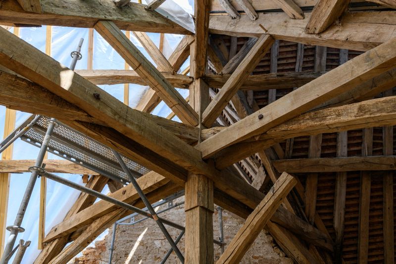 Detailed view of aged timber trusses and support beams in the roof structure of an old building under renovation. Natural light highlights the weathered wood, metal scaffolding, and textured masonry walls.