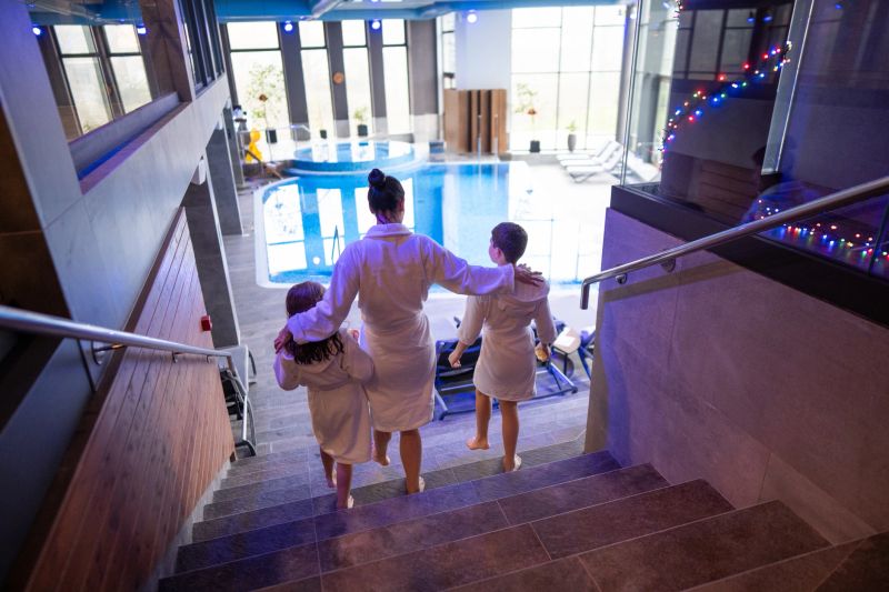 A family group in white robes walks down stairs to a bright indoor pool, creating a warm, relaxing spa vibe with festive string lights overhead.