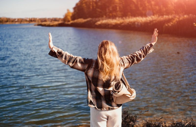 Woman standing by lake with raised arms, feeling freedom and relief, open autumn space, emotional release and mindful outdoor moment