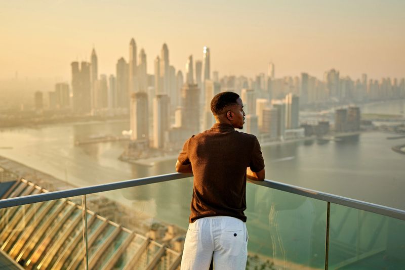Rear view of man leaning on railing, looking out at skyline with tall buildings, villas, marinas, and luxury in Dubai. Property release attached.