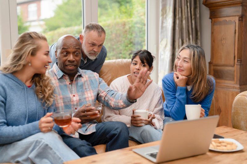 A group of five friends sit in a living room. They drink tea and coffee while sharing stories. Laughter fills the air as they engage in conversation and connect.