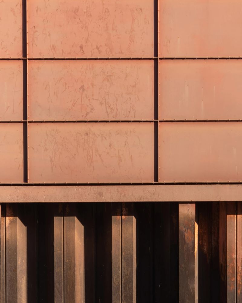Textured metal panels create a unique facade, reflecting warm afternoon sunlight in an urban environment. The design emphasizes modern aesthetics and industrial beauty.