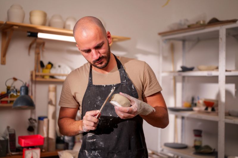 Ceramist smoothing a clay bowl with a specialized tool in a workshop, surrounded by shelves filled with various pottery pieces, showcasing the artistry and skill of handmade ceramics