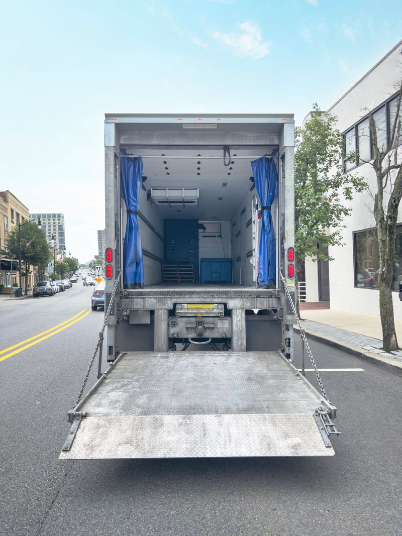 Rear view of an empty moving truck with its cargo door and loading ramp open, parked on a sunny city street ready for loading or unloading. Ideal for projects about relocation, delivery services, logistics, transportation, moving day and urban commerce.