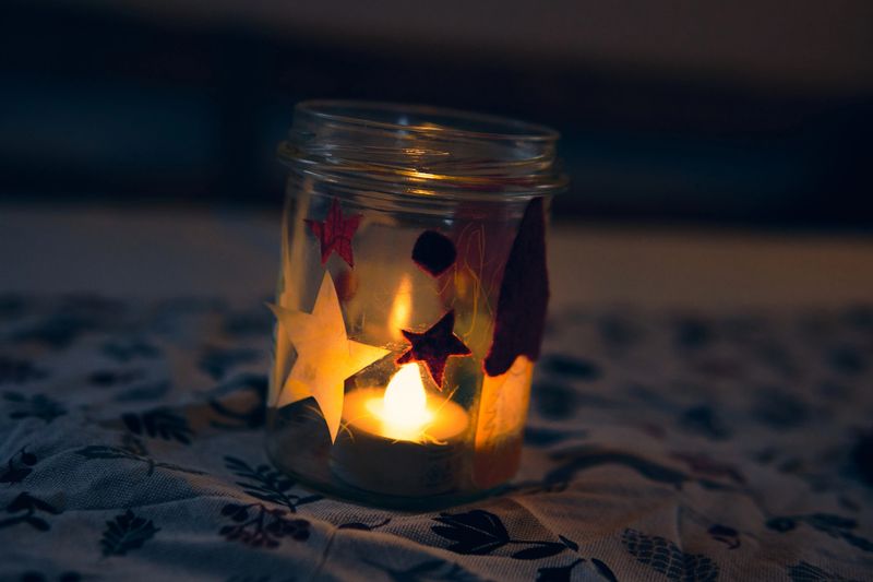 A candle is inside a jar decorated with colorful shapes. The jar sits on a patterned surface. The light from the candle creates a warm glow in the room.