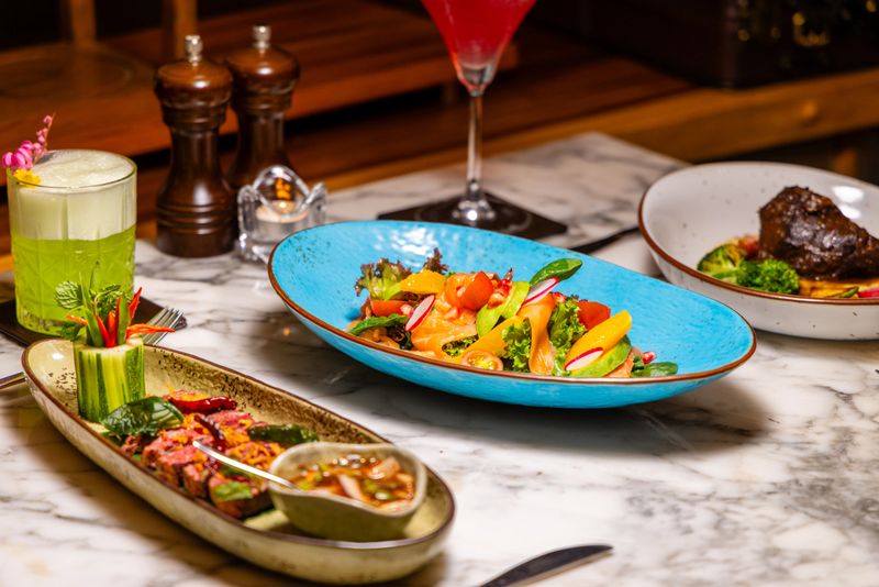 Gourmet dining experience with vibrant vegetable salad, Thai beef dish, and colorful cocktails on marble table in elegant restaurant setting.
