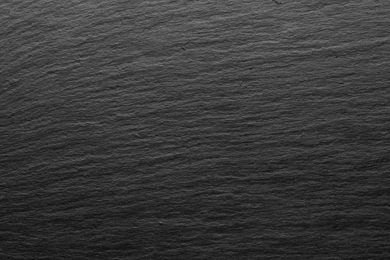 A high-resolution close-up of natural black slate stone with a rough, striated surface. The vertical grooves and matte finish give the image a raw, earthy quality, ideal for architectural, design, or background use.