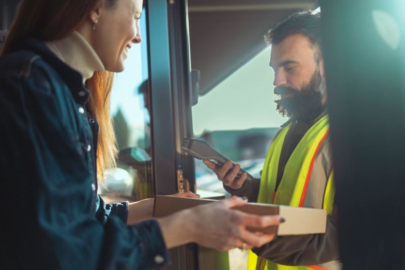 A delivery man in a high-visibility vest stands at the entrance of a home, handing a cardboard parcel to a female customer. He is holding a mobile POS terminal, ready for a contactless transaction