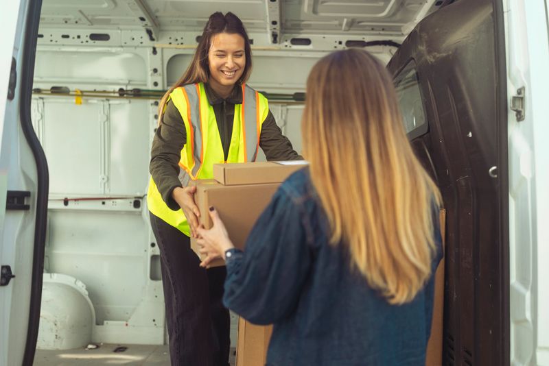 Female delivery courier wearing a reflective safety vest smiles while handing a package from the delivery van's trunk to a female customer