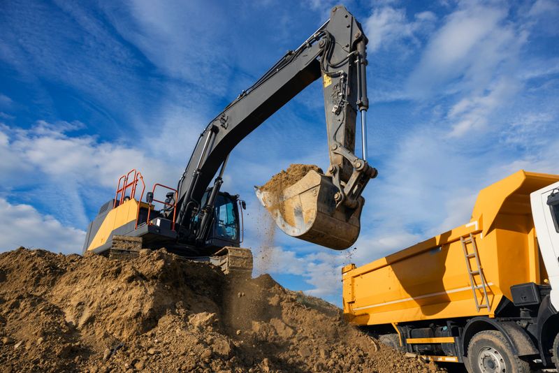 Heavy excavator loading soil into a dump truck at an active construction site under a blue sky. This image represents excavation work, earthmoving operations, infrastructure development, and the construction industry. Ideal for concepts such as building projects, heavy machinery, industrial work, civil engineering, road construction, and urban development.