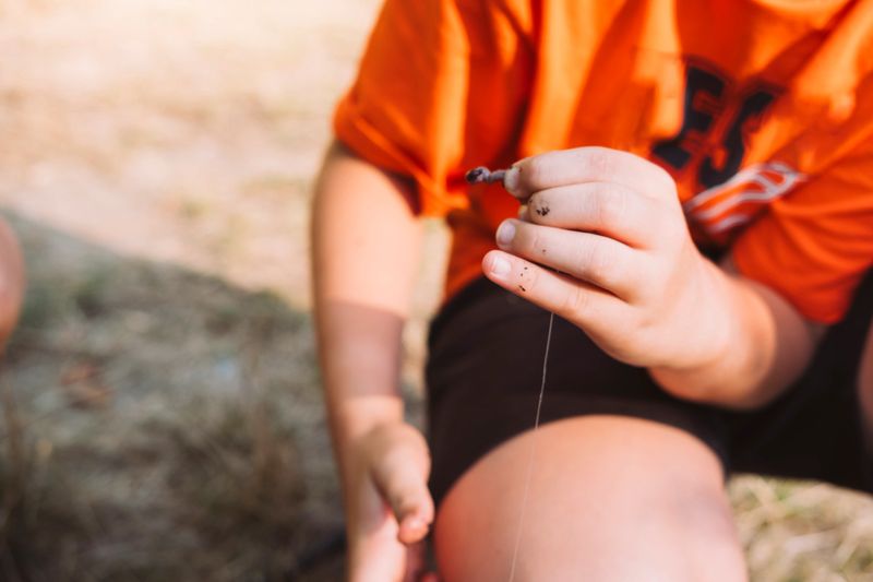 A boy in an orange shirt holding a worm as fishing bait in his hand, preparing for fishing.