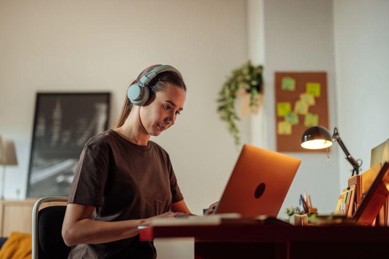 A focused woman sits at a desk with a laptop and headphones, creating a calm, productive work atmosphere in a modern home office with soft lighting, plants, and organized supplies.