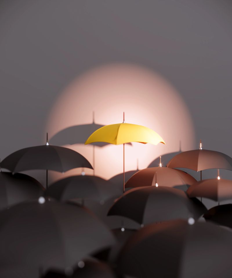 A single yellow umbrella is highlighted by a soft spotlight among dark umbrellas, symbolizing individuality, confidence, and positive risk-taking within a crowd. Minimal studio lighting and clean composition emphasize focus and contrast. (3D render)