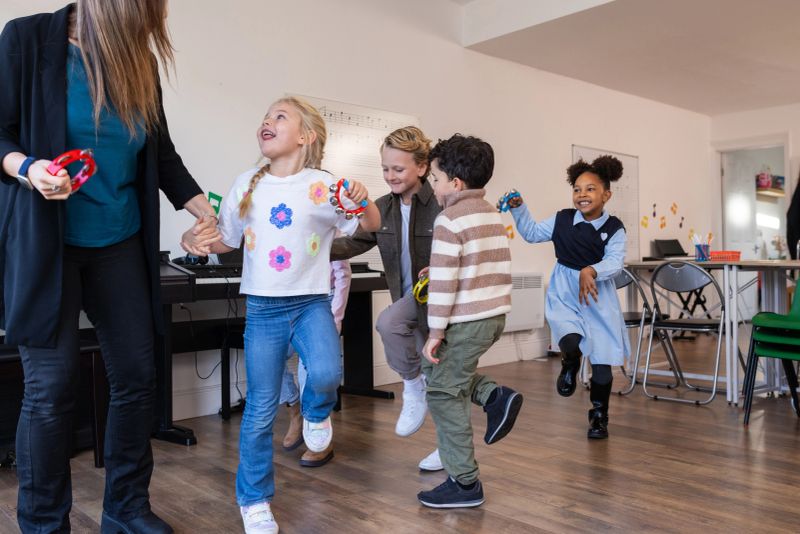 A group of excited children and their tutor are standing and dancing in a bright classroom, playing colourful percussion instruments during a music lesson. The scene represents early childhood education, creativity, teamwork, and musical development in a preschool setting.Video for this scenario is available