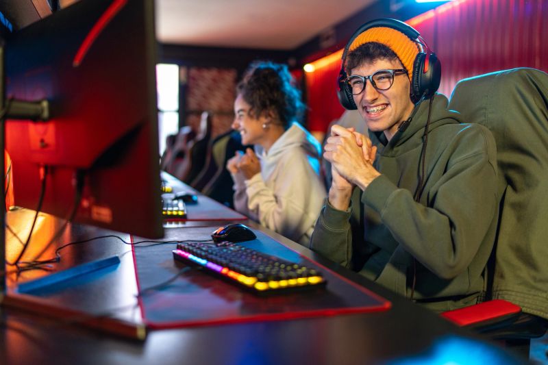 Happy young man wearing headphones and glasses with braces celebrating a win while playing competitive video games at an e-sports game center with friends