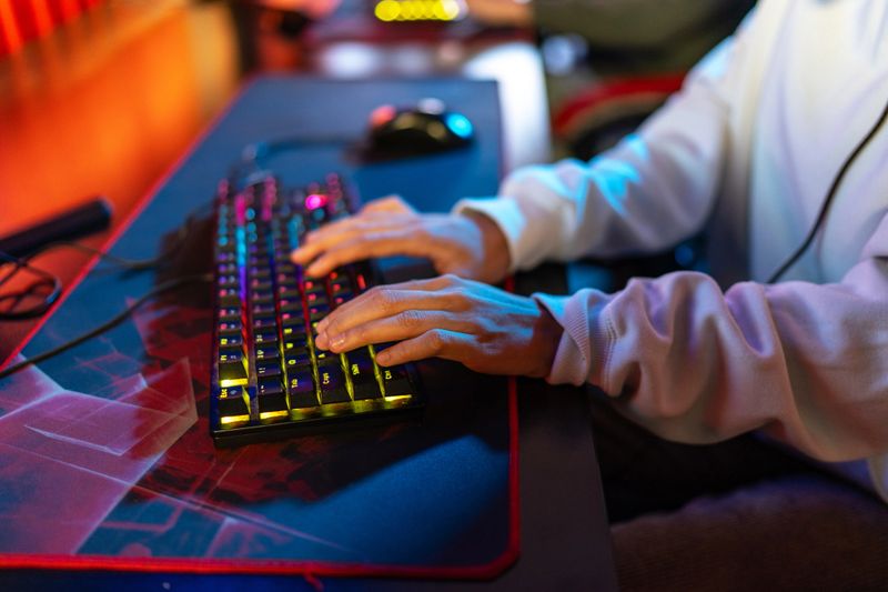 Unrecognizable gamer hands typing on a mechanical rgb keyboard with illuminated keys on a gaming mouse pad, enjoying an online computer game during a competitive esport event
