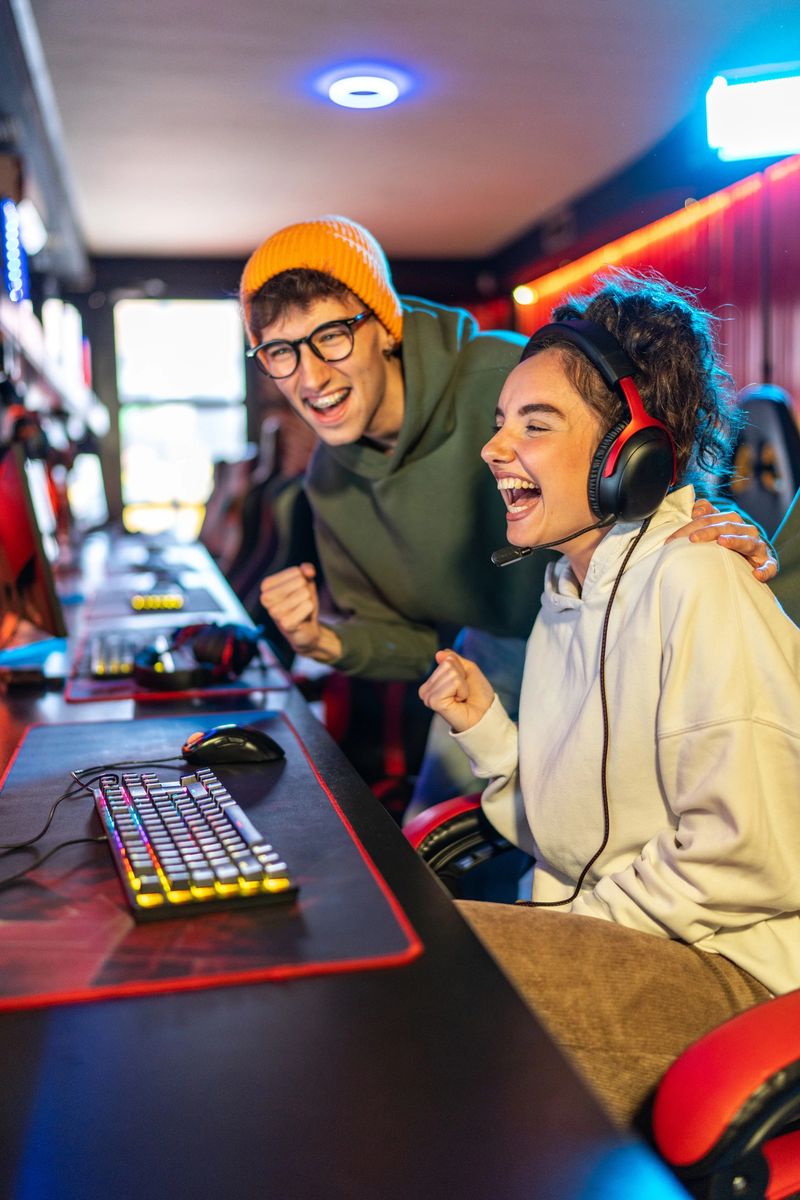 Excited young adult friends cheer and fist pump at a neon-lit esports station, celebrating a competitive win together while streaming and gaming in a lively arena lounge