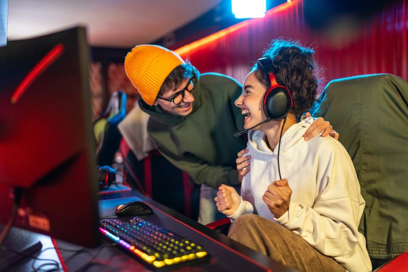 Two young adult gamers wearing headsets, smiling and celebrating a thrilling online victory in a neon-lit gaming room while streaming and high-fiving over their keyboards and monitors
