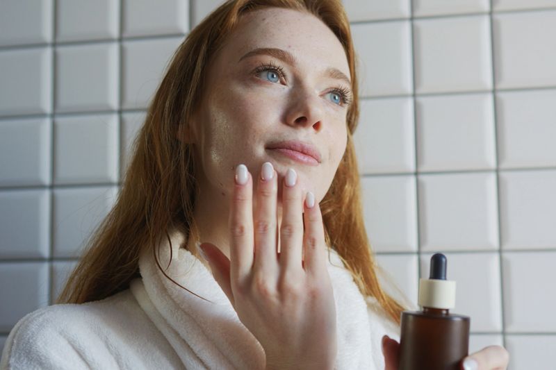 Woman applying hydrating serum with dropper and bottle, careful fingertip application on chin, tiled bathroom backdrop, focused