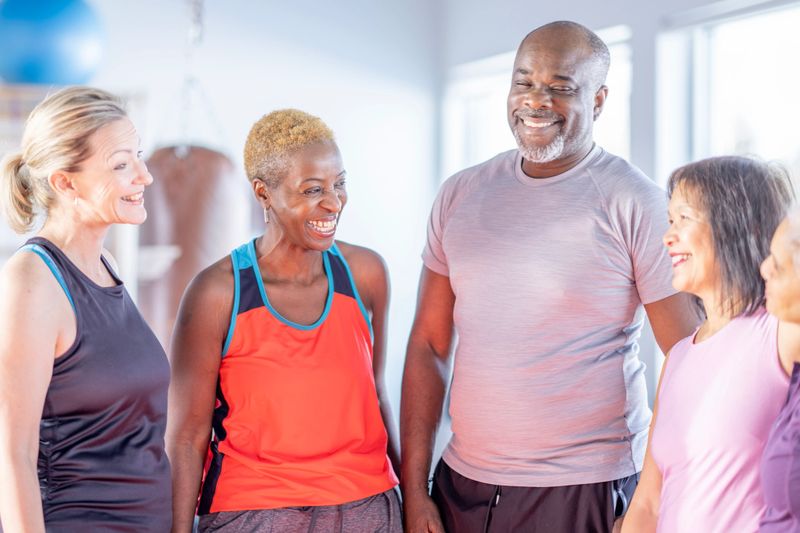 A diverse group of adults in a bright gym sharing a joyful moment. They chat and smile during a fitness class, showcasing camaraderie, health, and community.