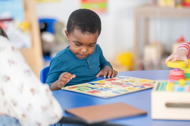 A young African American child focuses on a colorful puzzle at a blue table in a bright, playful learning environment. Educational toys and stacking blocks are visible nearby, highlighting early learning and exploration.