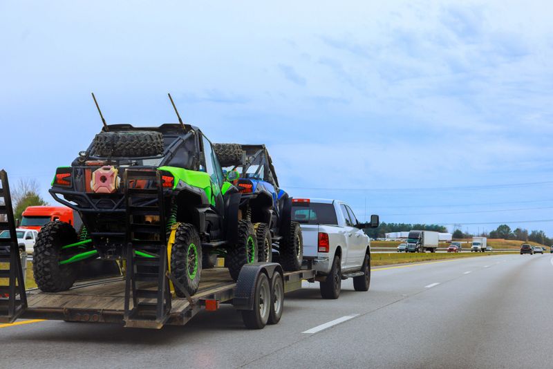 Truck pulls trailer with two off road vehicles down busy American highway with other vehicles are nearby.