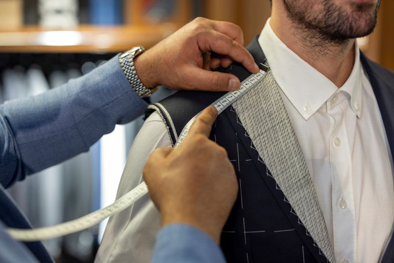 Close-up of a professional tailor measuring shoulder width of a bespoke suit during a fitting session. Concept of custom tailoring, menswear, precision, luxury fashion, craftsmanship and personalized clothing service.