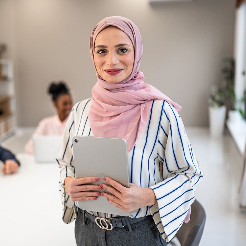 Confident muslim businesswoman smiling at camera, holding a digital tablet in a modern diverse office workplace