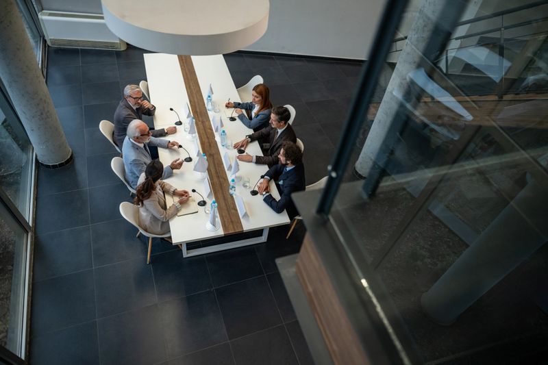 Diverse group of diplomats and executives at a modern conference table, engaged in serious international negotiation and policy discussion with microphones and documents