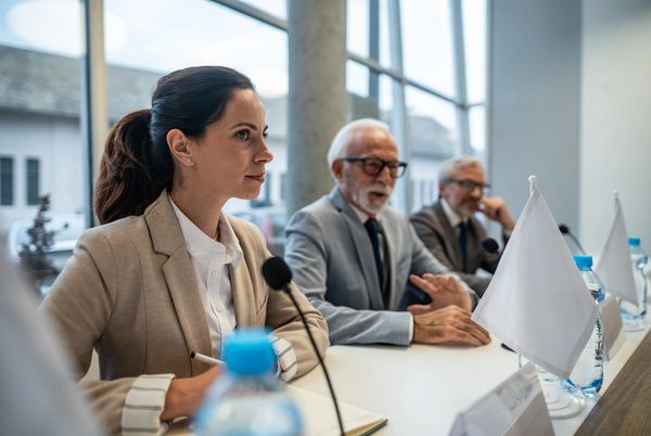 A professional woman and two men at a conference table with microphones and water bottles.